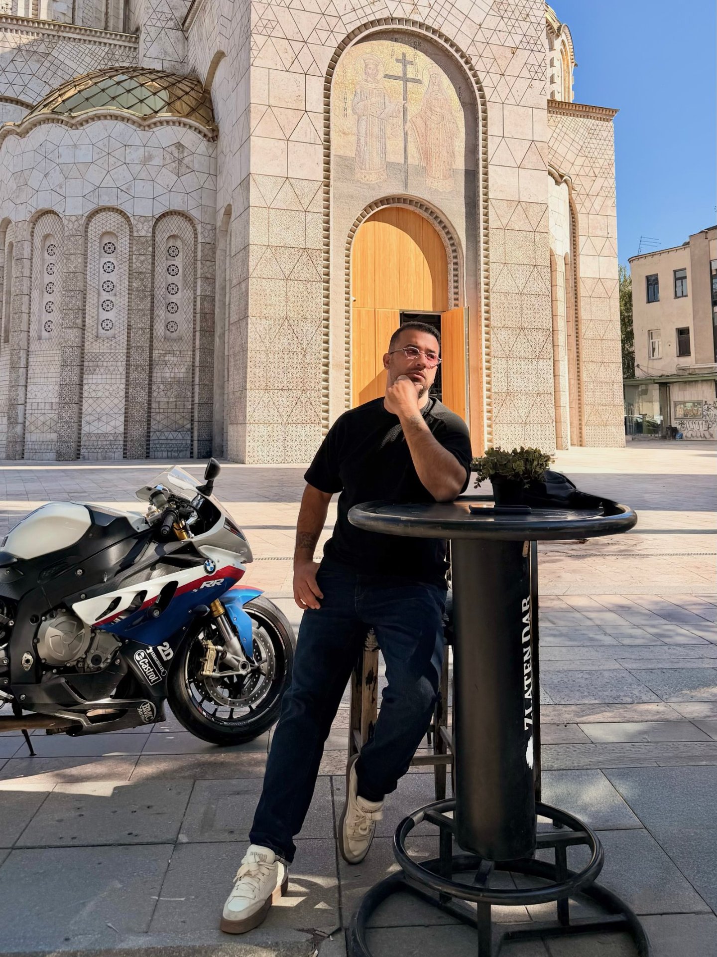 Batuhan Emir Alkan, drone videographer and photographer, leaning on a high table in front of a stone church next to a BMW S1000RR